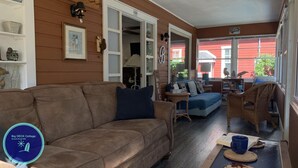 A cozy living room with a brown couch, a blue sofa, and wicker chairs. There is a bookshelf with books and decorative items. A sign on the wall reads "BIG DECK Cottage." - Big DECK Cottage, 3-min walk to BEACH- 4 bdrms, 1.5 baths 7 beds, parking 3 cars (Grand Bend)
