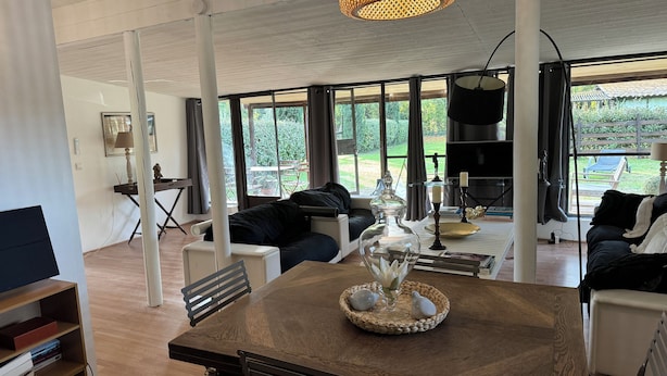 A spacious living room with a large wooden table, black sofas, and a television.