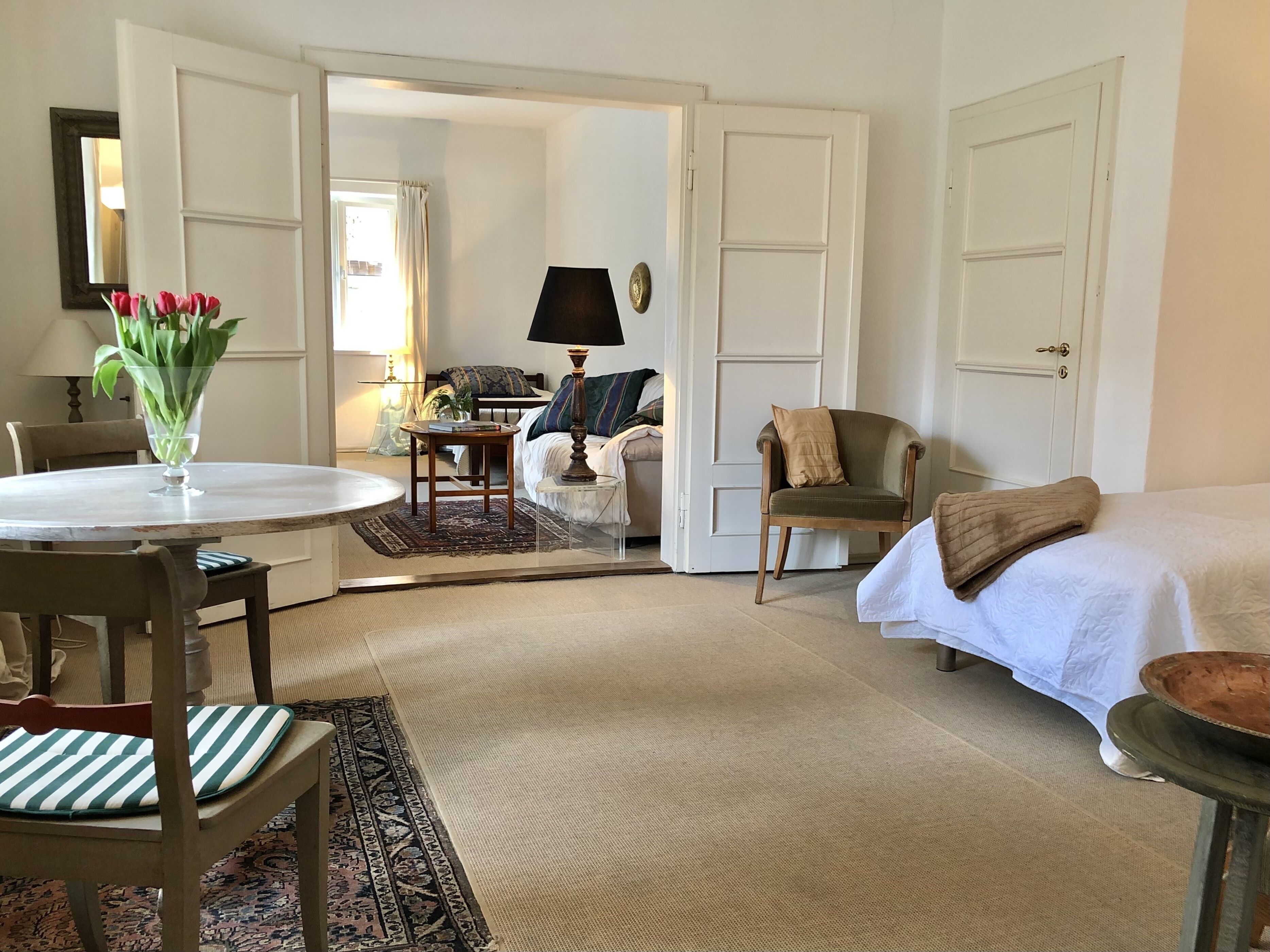 Ein Zimmer mit einem Bett, einem Sessel, einem Tisch mit Blumen und einer Lampe, sowie einer Tür zu einem anderen Raum.