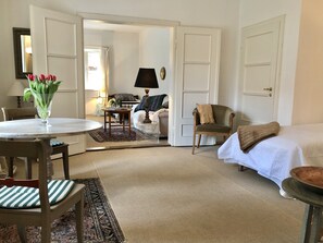 Ein Zimmer mit einem Bett, einem Sessel, einem Tisch mit Blumen und einer Lampe, sowie einer Tür zu einem anderen Raum.