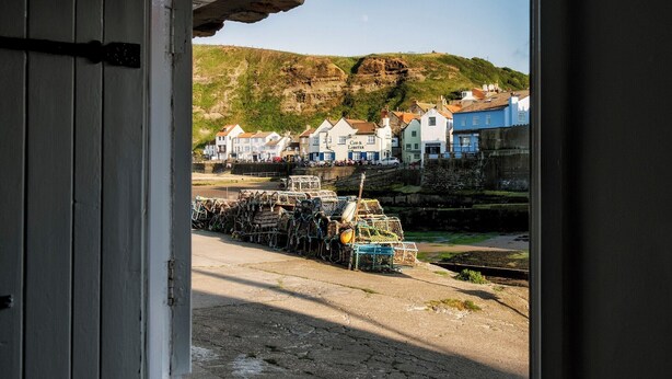 A coastal town with buildings along the waterfront, a dock with lobster traps, and a rocky hill in the background. - A delightful fisherman’s cottage with uninterrupted sea views. (Staithes near Whitby)
