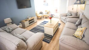 A living room with a flat-screen TV mounted on a blue wall, a coffee table with a vase of flowers, and two sofas with patterned cushions.