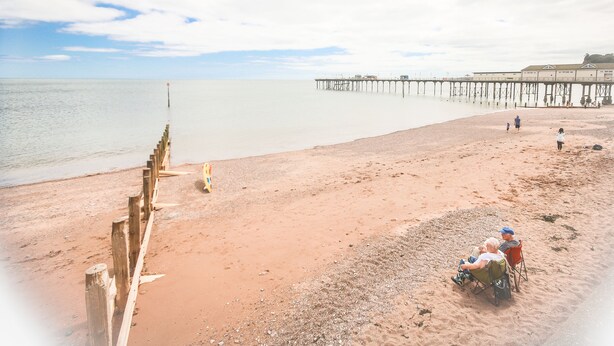 A beach with a wooden jetty extending into the water, a clear sky, and a few people walking.