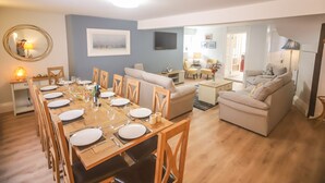 A dining area with a long wooden table set for six, surrounded by wooden chairs. There is a sofa and a small table with a lamp in the living area.