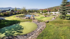Un campo de golf con un estanque, rodeado de árboles y montañas al fondo.