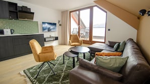 Ein modernes Wohnzimmer mit einem braunen Ledersofa, zwei Sesseln, einem Couchtisch und einer Küchenzeile mit dunklen Schränken und einer grünen Spritzschutzwand.
