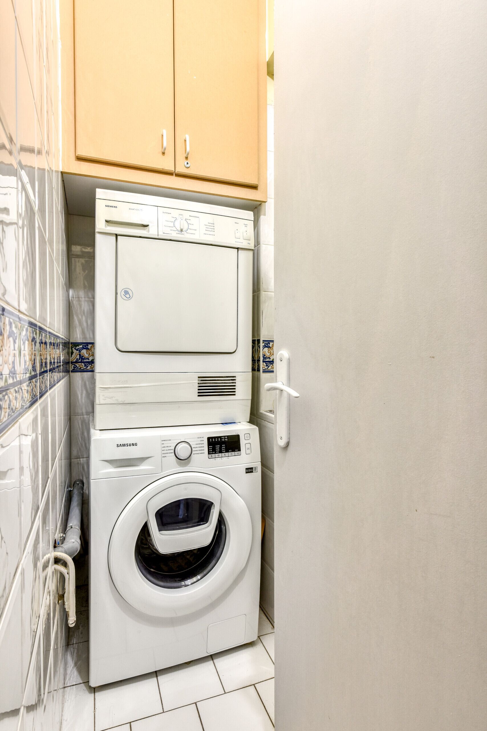 Une petite buanderie équipée d’un lave-linge et d’un sèche-linge superposés, d’un pare-feu en carrelage et d’armoires en bois.