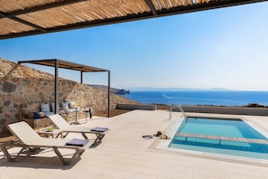 Un espace piscine extérieur avec des chaises longues, une petite table et une pergola offrant une vue sur la mer.
