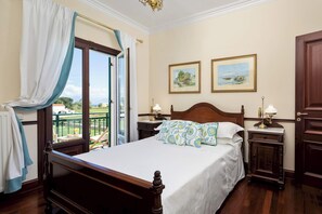 A bedroom with a wooden bed, two bedside tables with lamps, a large window with curtains, and two framed pictures on the wall.