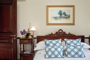 A neatly made bed with white linens and two blue and white checkered pillows, a wooden nightstand with a lamp and a vase of purple flowers, and a framed landscape painting on the wall.