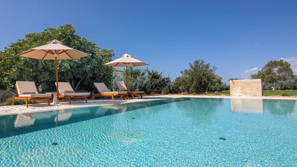 Piscina azul cristalina com espreguiçadeiras e guarda-sóis, rodeada por vegetação.