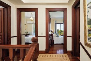 A hallway with wooden doors leading to a bedroom with a bed, a nightstand, and a dresser. There is a chandelier hanging from the ceiling.