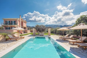A pool area with lounge chairs and umbrellas, a pergola, and a view of mountains.