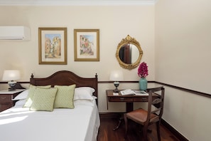 A hotel room with a bed, two framed pictures, a mirror, a desk with a lamp, a chair, and a vase with flowers.