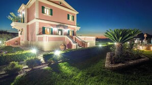 A two-story house with a balcony, a staircase, and a well-lit garden area at night. - Villa Rose Corfu, Villa (Acharavi)