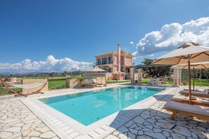 A swimming pool with lounge chairs and a hammock, surrounded by stone tiles and a large house in the background.