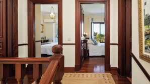Um corredor com portas de madeira que levam a um quarto com cama, mesa de cabeceira e cômoda. Um lustre pendurado no teto.