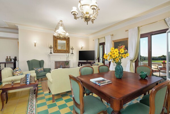 A spacious living room with a large wooden dining table, green upholstered chairs, a fireplace, a television, and a large window with curtains.