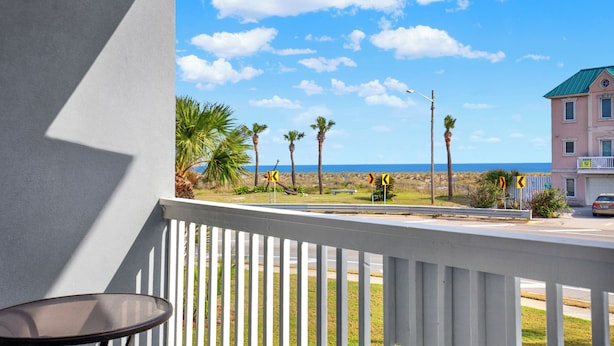 A balcony with a view of the ocean, palm trees, and a pink building.