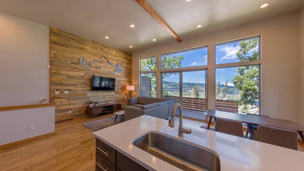 A modern kitchen with a large island, wooden wall paneling, a flat-screen TV, and a dining area with a view of the outdoors. - Hawks Landing - New, Modern, Views, Billiards! (Hood River)