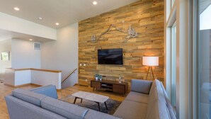 A modern living room with a wooden wall, a flat-screen TV, a grey sofa, a coffee table, and a floor lamp. - Hawks Landing - New, Modern, Views, Billiards! (Hood River)