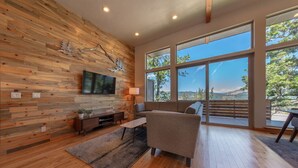 A modern living room with a wooden wall, a flat-screen TV, a sofa, and a large window offering a scenic view. - Hawks Landing - New, Modern, Views, Billiards! (Hood River)