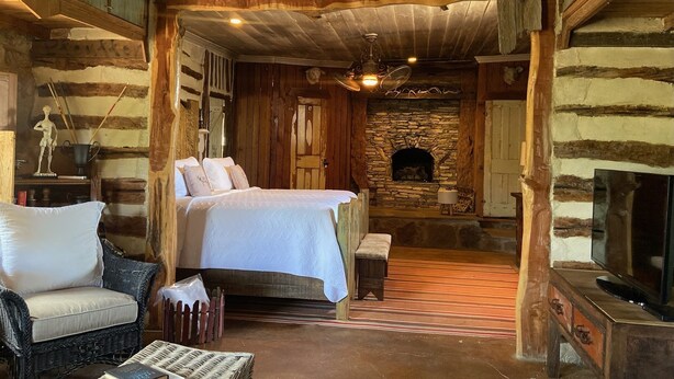 A rustic log cabin bedroom with a stone fireplace, a bed, a wicker chair, and a television. - Stay at these upscale, historic cabins set in New Braunfels near Guadalupe River (New Braunfels)