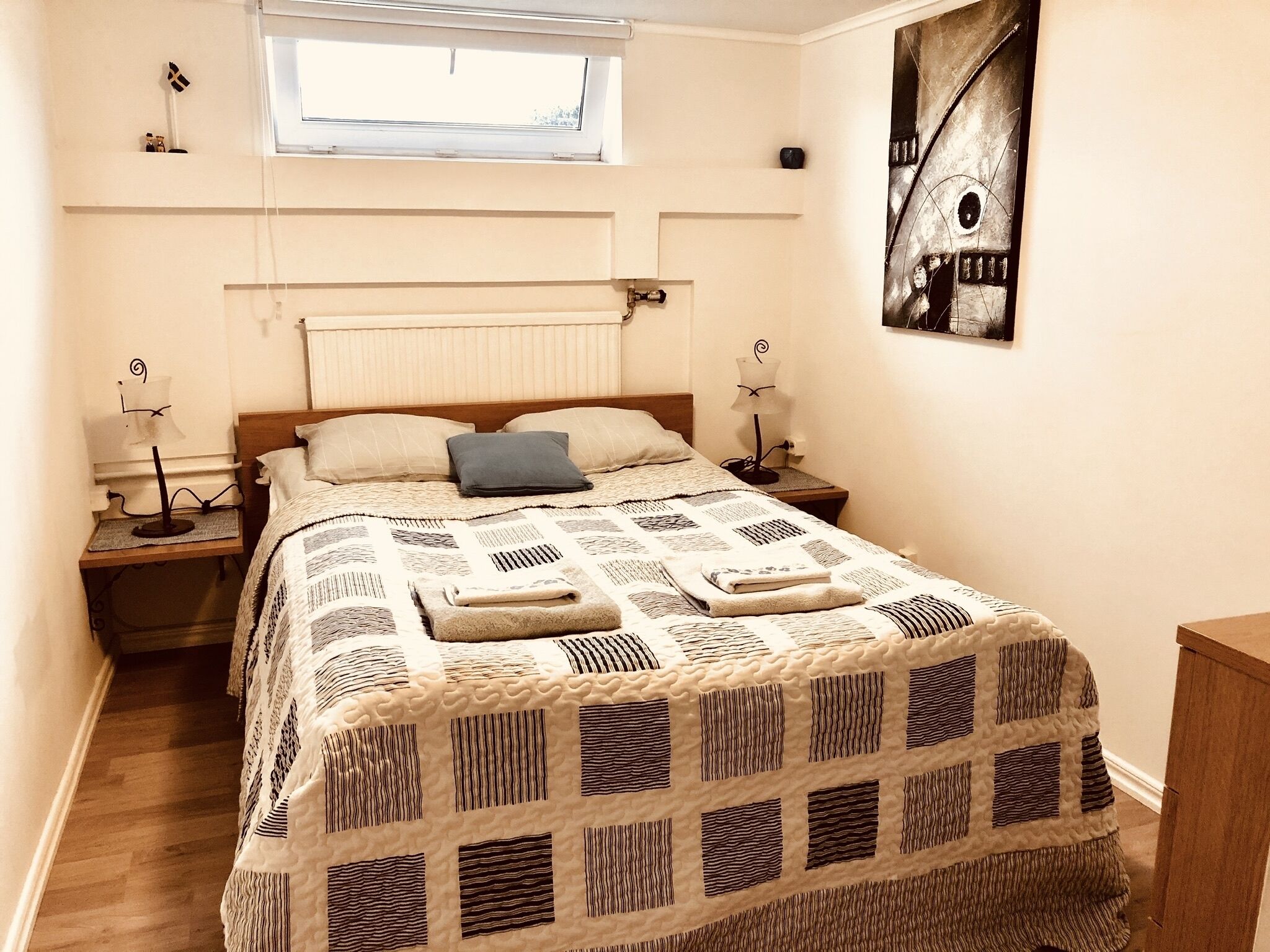 A neatly made bed with a patterned bedspread, two bedside lamps, a window with blinds, and a wall-mounted artwork.