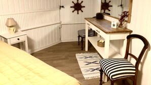 A compact bedroom with a bed, a bedside table with a lamp, a wooden dresser, a chair, a window, and a mirror.