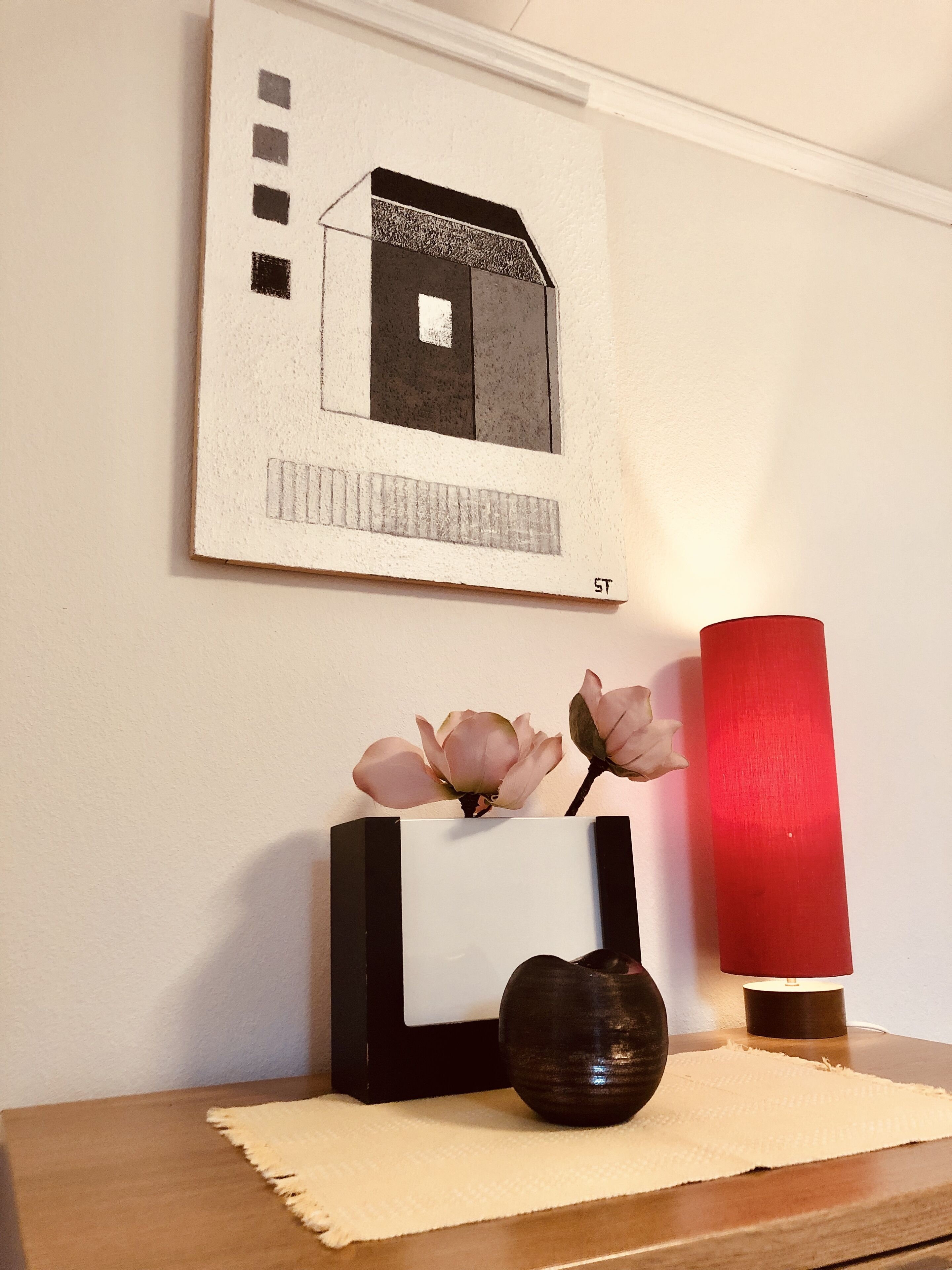 A framed artwork of a house on a wall, a vase with flowers, and a red candle holder on a table.