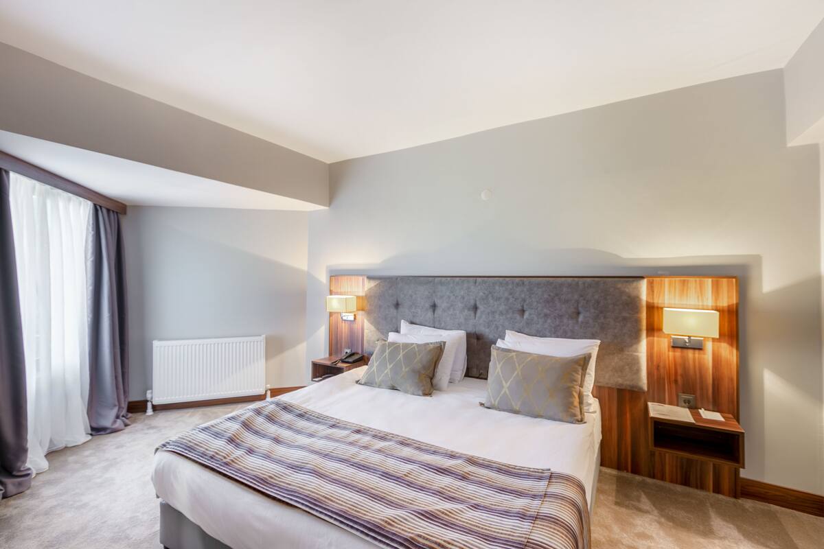 A modern hotel bedroom with a large bed, two bedside tables with lamps, a radiator, and a window with curtains.