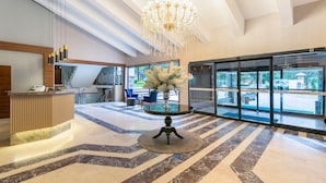Un hall d’hôtel comprenant un comptoir d’accueil, un lustre, une table ronde ornée d’un vase de fleurs et donnant sur l’extérieur grâce à de grandes portes vitrées.