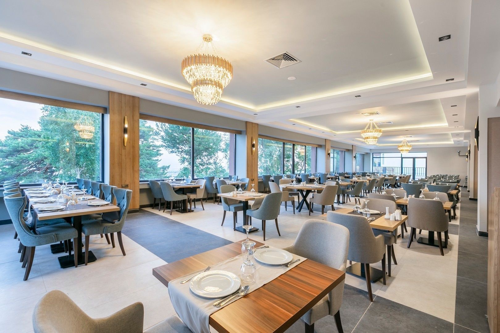 A modern dining area with multiple tables and chairs, large windows offering a view of trees, and elegant chandeliers.