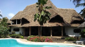 Un resort con una construcción de techo de paja, piscina y jardines tropicales.