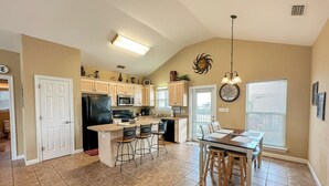 Una cocina amplia con una isla central, un área de comedor con una mesa y sillas de madera, y un ventanal con persianas.