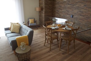 A modern living room with a grey sofa, yellow cushions, a round wooden dining table with chairs, and a brick-patterned wall.