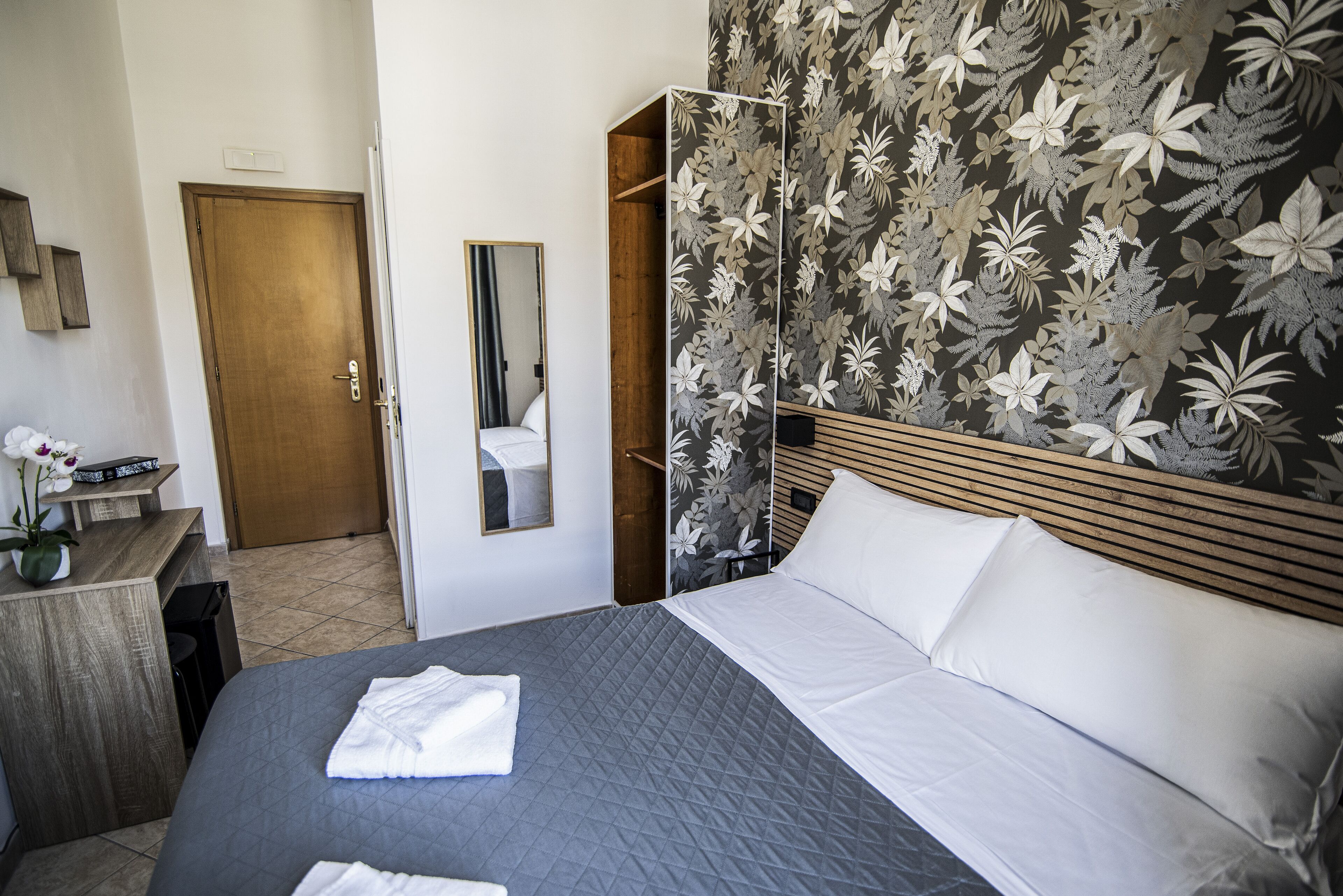 Una camera d'albergo con un letto, una testiera in legno, uno specchio e carta da parati con motivi floreali.