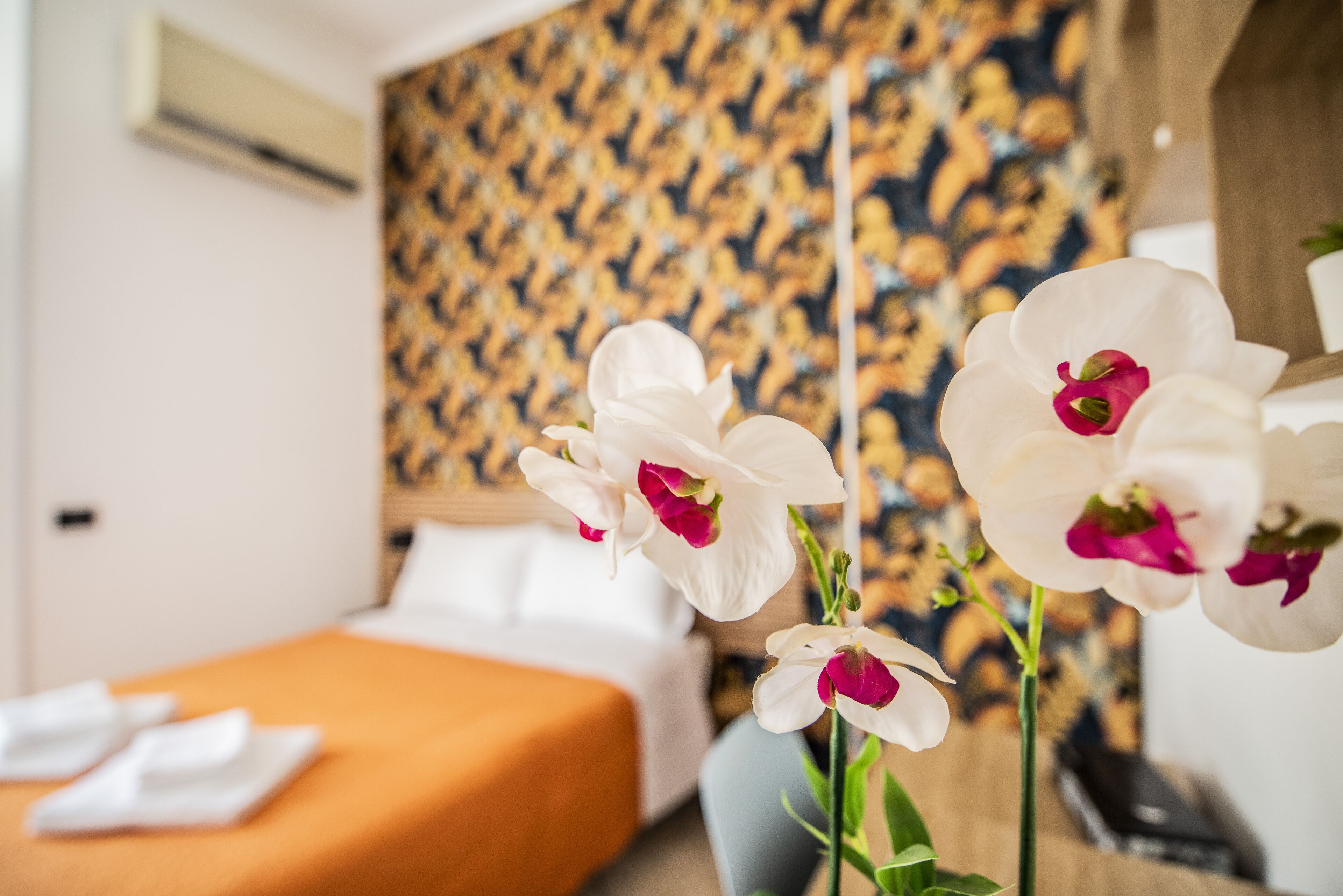 A hotel room with a bed, an orange bedspread, white pillows, and white orchids.