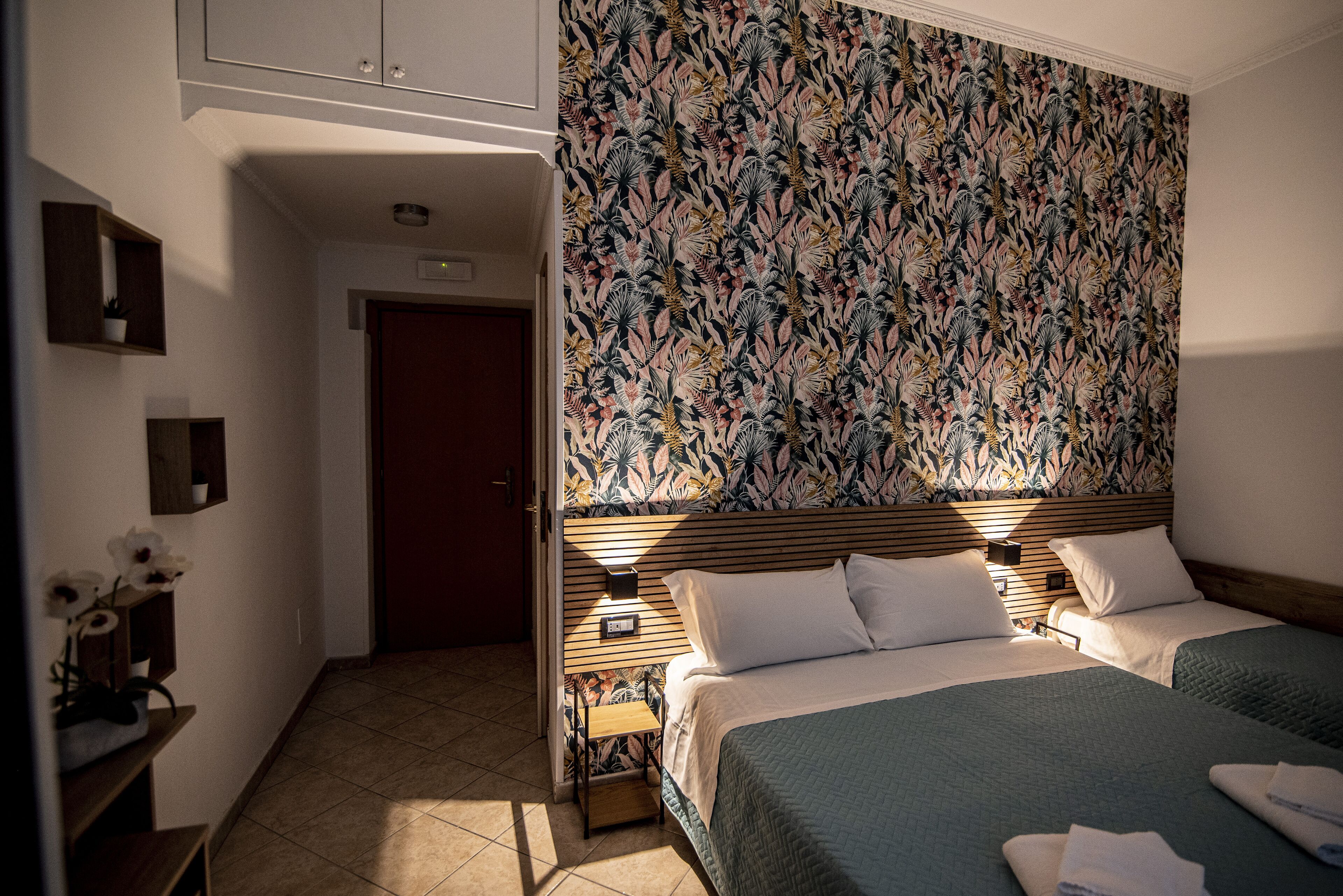 Una habitación de hotel con dos camas, cabecera tapizada con estampado floral, mesita de noche de madera y una pequeña estantería con planta en maceta.