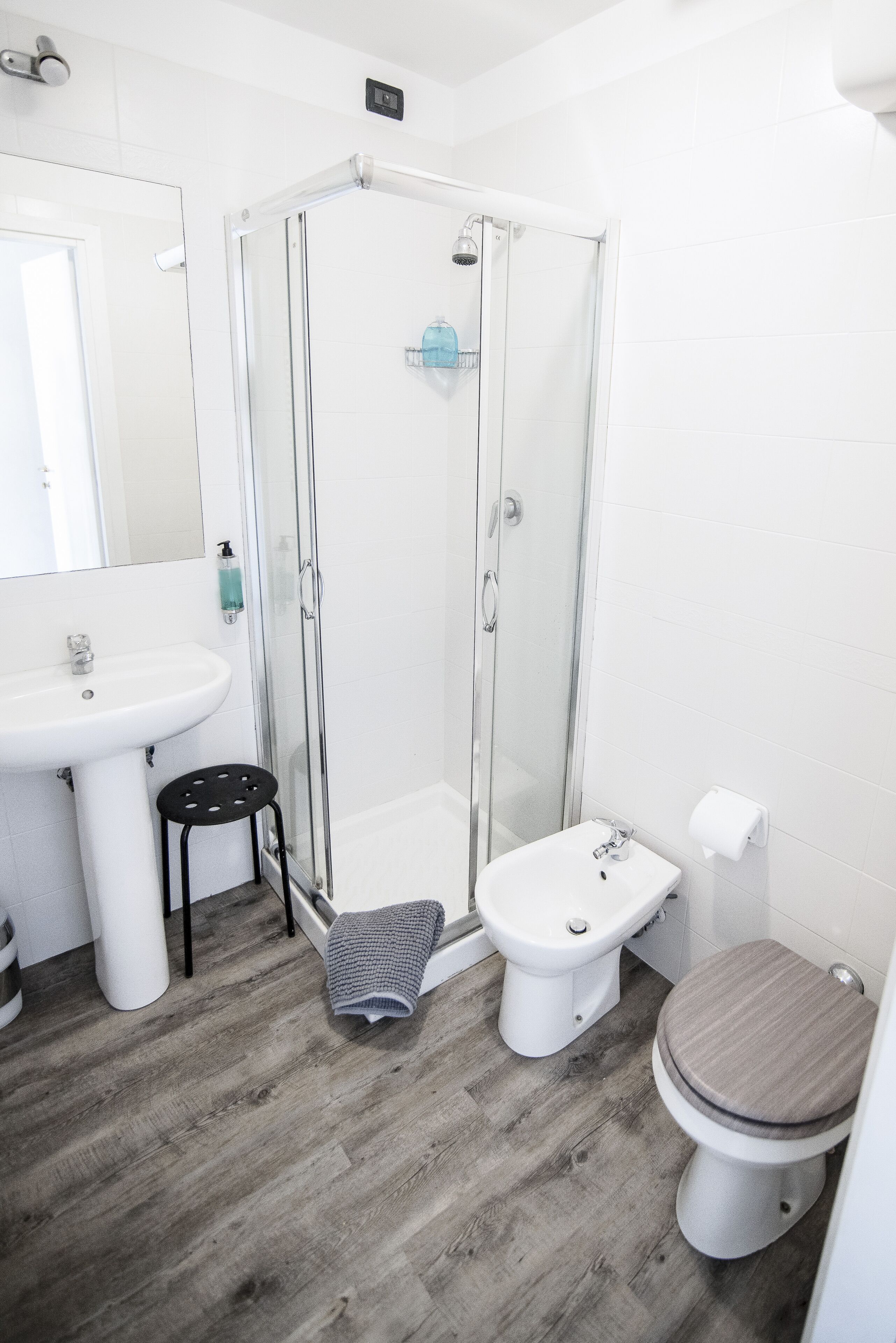 Un baño moderno con cabina de ducha de vidrio, lavamanos pedestal, inodoro y suelo de madera.