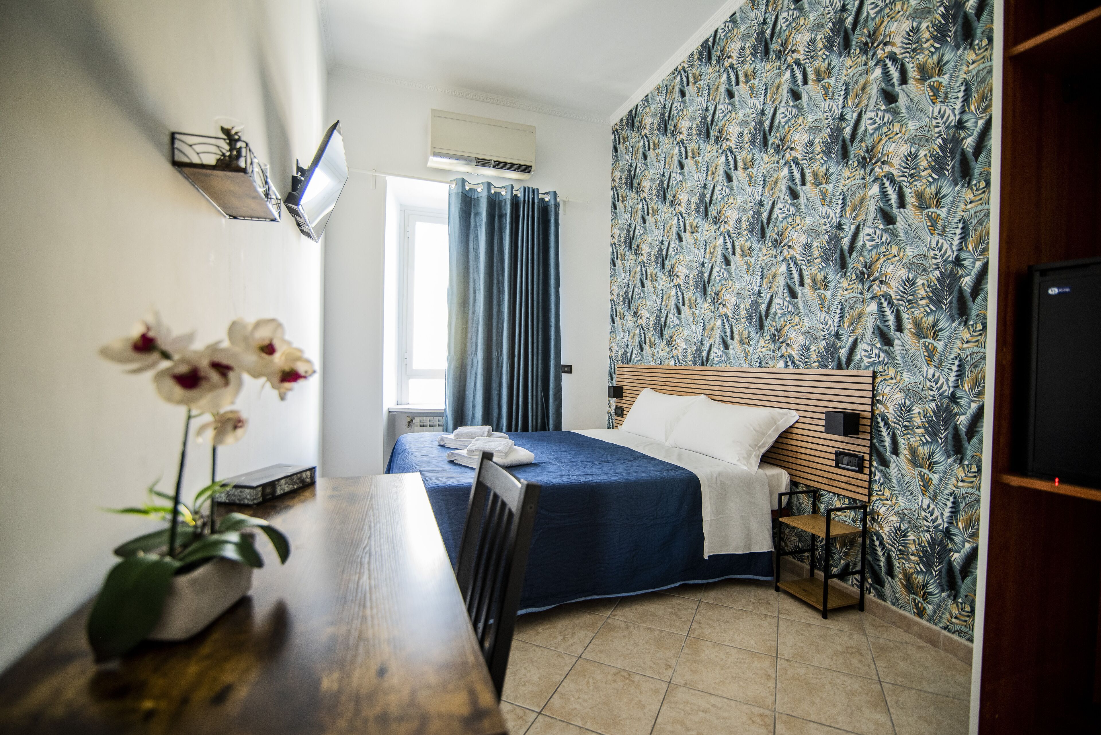 Ein Hotelzimmer mit einem Bett, einem Nachttisch aus Holz, einem Schreibtisch mit Stuhl, einer Topfpflanze und einem an der Wand montierten Fernseher.