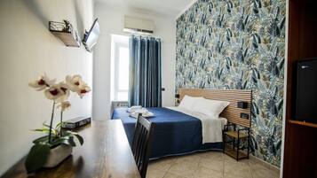 Ein Hotelzimmer mit einem Bett, einem Nachttisch aus Holz, einem Schreibtisch mit Stuhl, einer Topfpflanze und einem an der Wand montierten Fernseher.