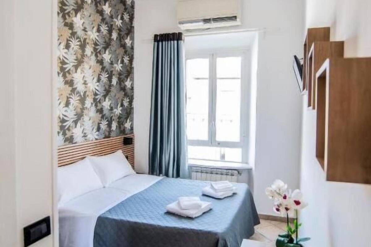 A hotel room with a bed, a desk, a window with curtains, a wall with a floral pattern, and a ceiling light.