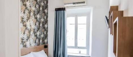 A hotel room with a bed, a desk, a window with curtains, a wall with a floral pattern, and a ceiling light.