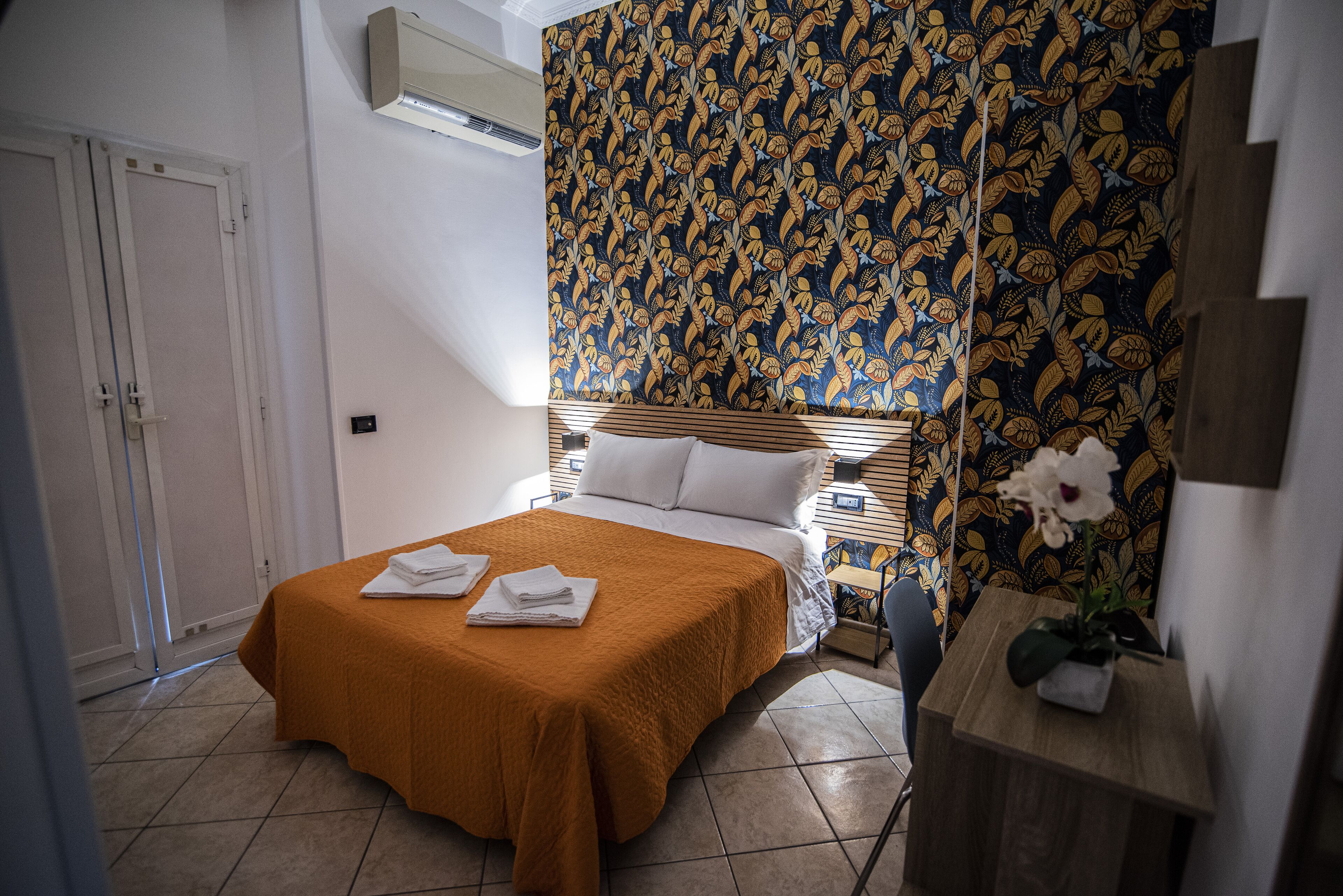 Une chambre à coucher comprenant un lit, une table de chevet, une petite table en bois, un papier peint à motifs et une porte.