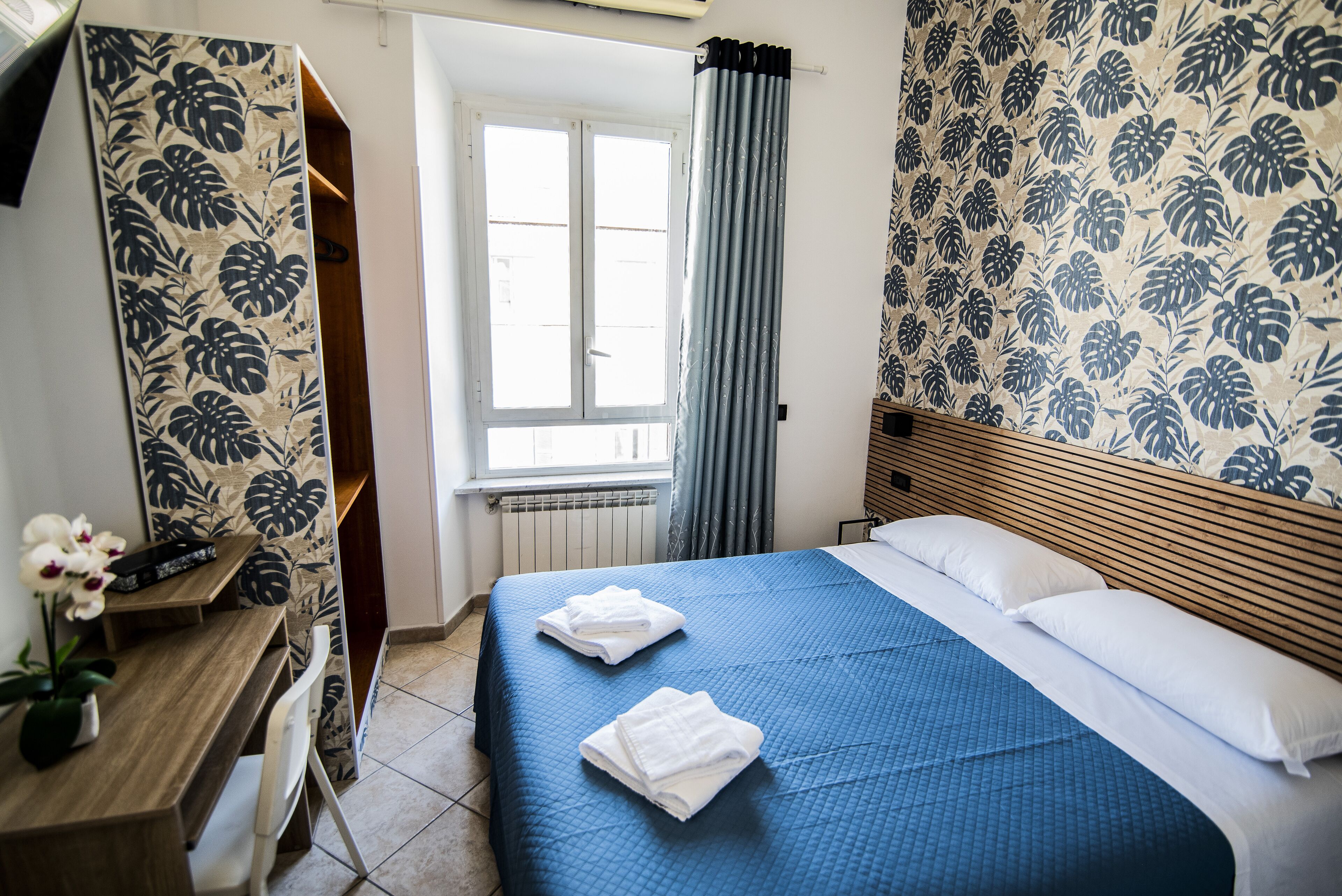 A hotel room with a bed, a desk, a chair, a window with curtains, and a wall with a floral pattern.