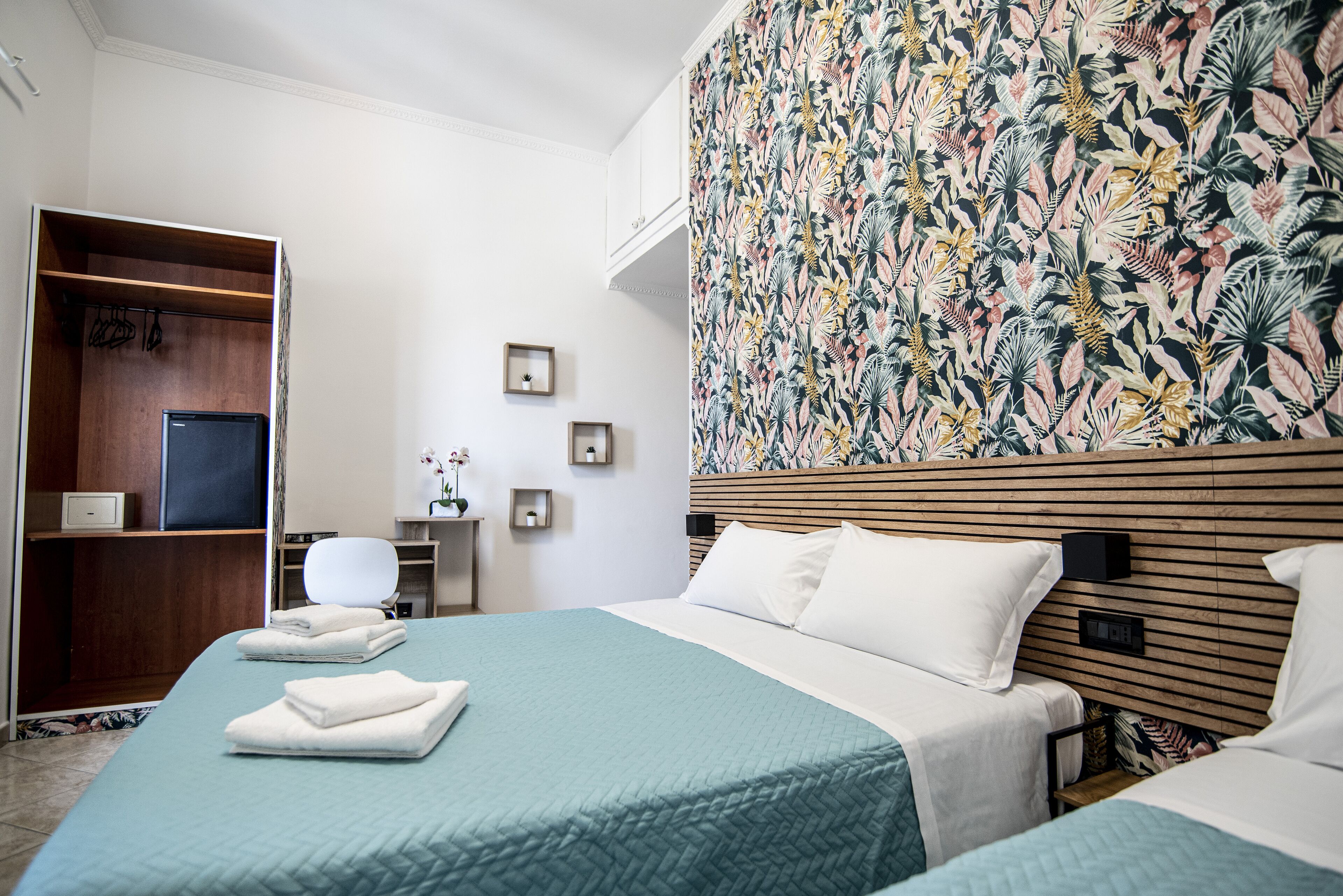 A hotel room with a bed, a wardrobe, a desk, and a wall with a floral pattern.