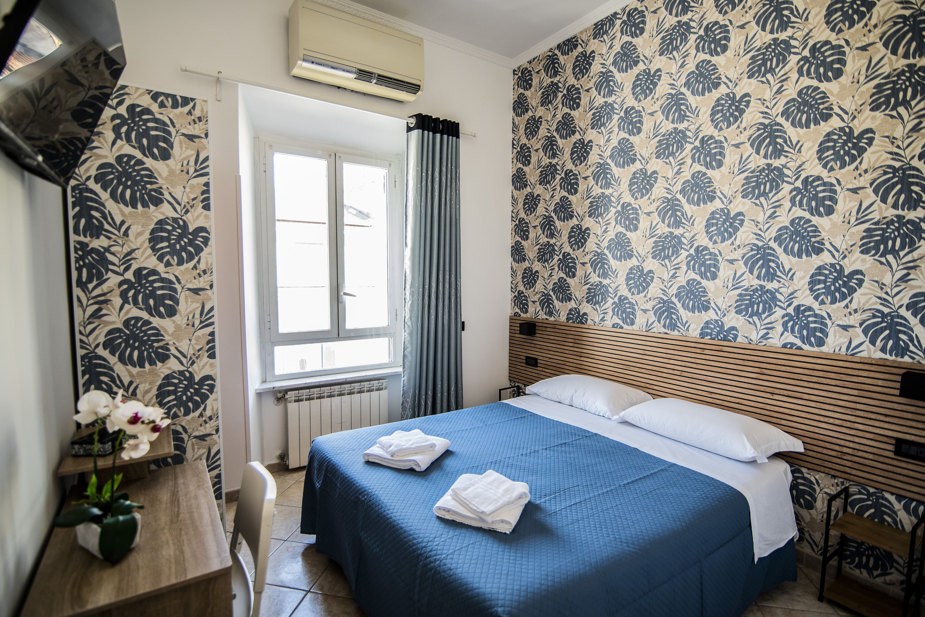 A bedroom with a bed, a window with curtains, a radiator, a wooden headboard, and a wall with a floral pattern.