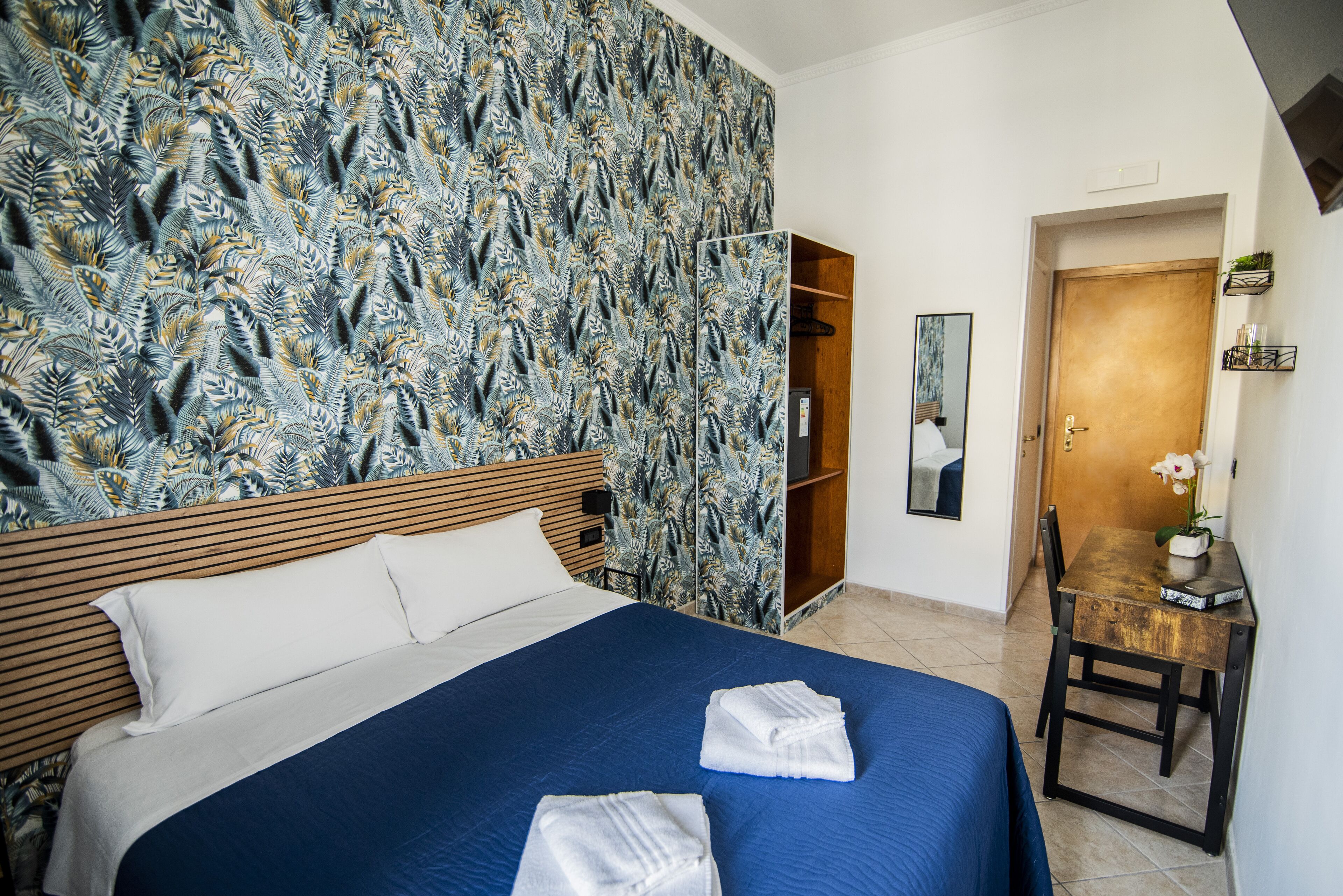 A hotel room with a bed, a wooden headboard, a blue bedspread, and a patterned wallpaper.