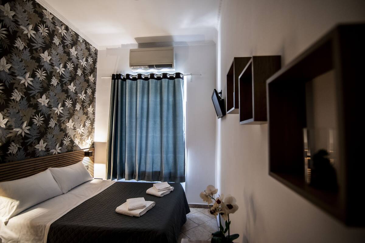 A hotel room with a bed, a window with curtains, an air conditioning unit, and a wall-mounted TV.